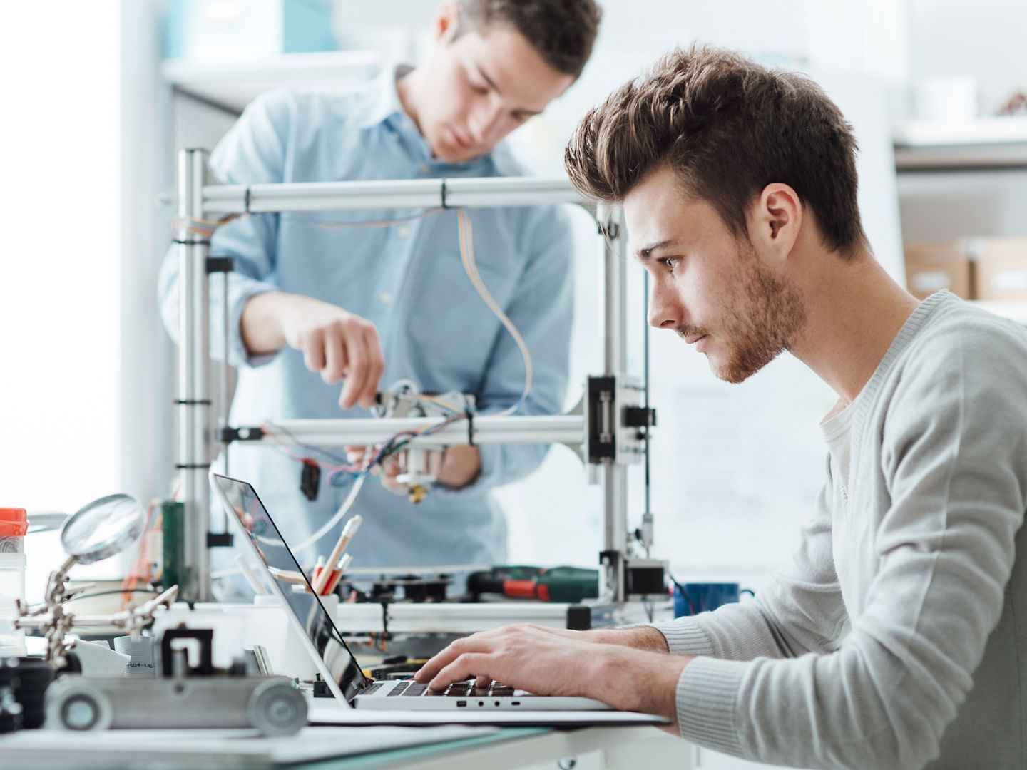 Engineering students working in the lab, a student is adjusting a 3D printer's components, the other one on foreground is using a laptop