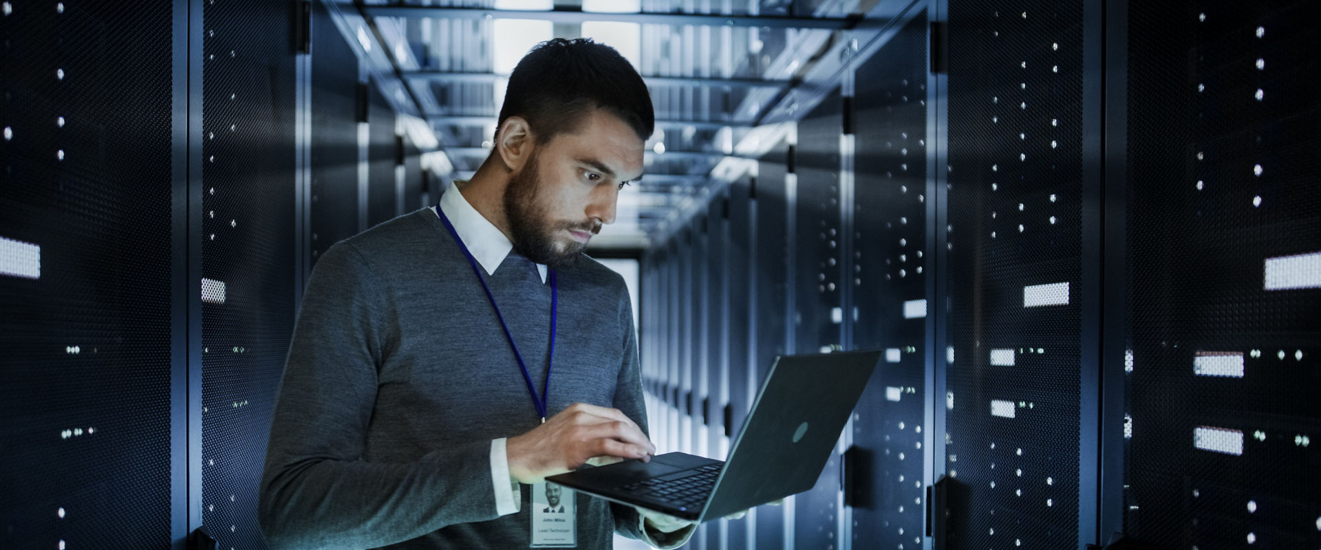 IT Technician Works on a Laptop in Big Data Center full of Rack Servers. He Runs Diagnostics and Maintenance, Sets up System.