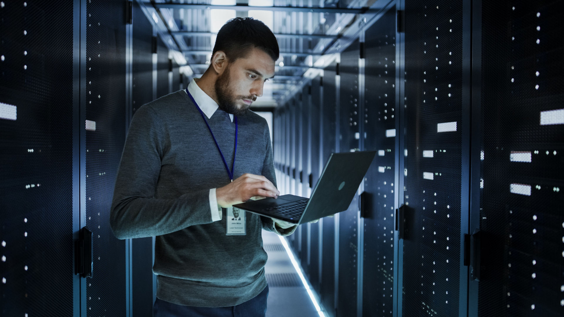 IT Technician Works on a Laptop in Big Data Center full of Rack Servers. He Runs Diagnostics and Maintenance, Sets up System.