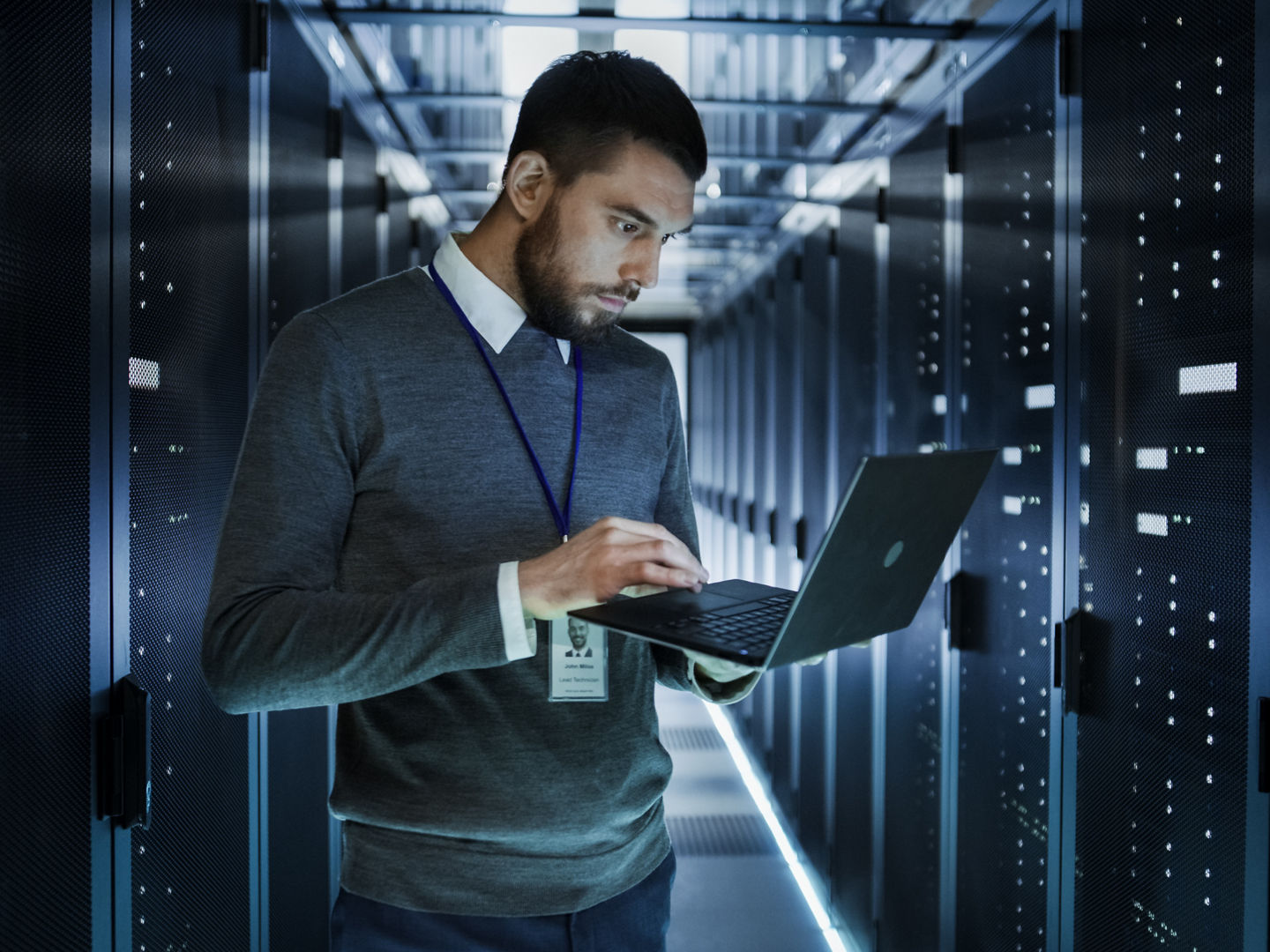 IT Technician Works on a Laptop in Big Data Center full of Rack Servers. He Runs Diagnostics and Maintenance, Sets up System.