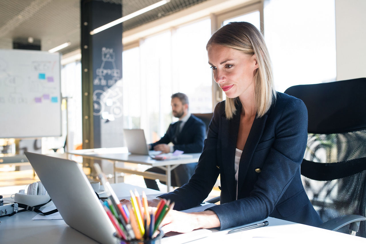 Two attractive young business people with laptops in the office, sitting at the desks, working.