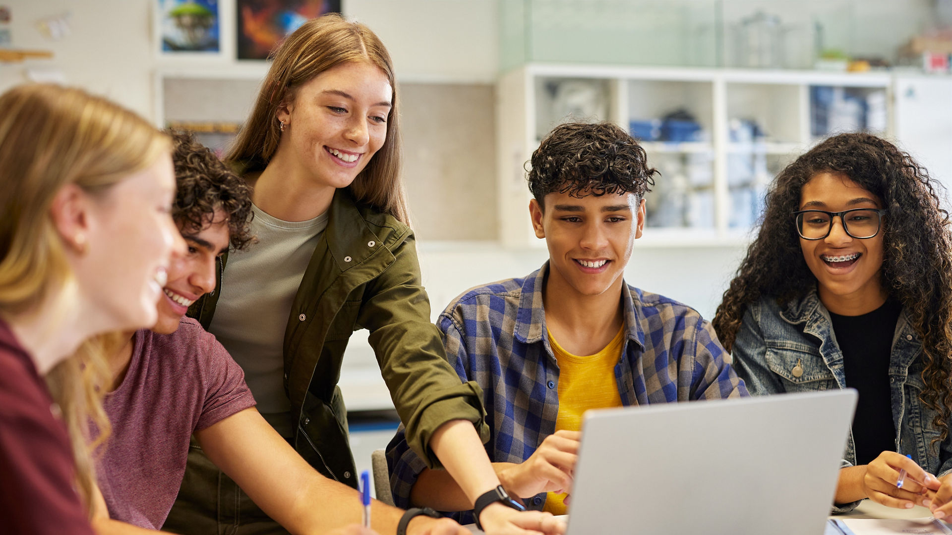 Multiethnic casual students using laptop during research, working together on a project. Diverse teenagers pointing at screen while engaging in collaborative learning. Group of happy classmates sharing ideas and taking notes during a digital study session for exam.