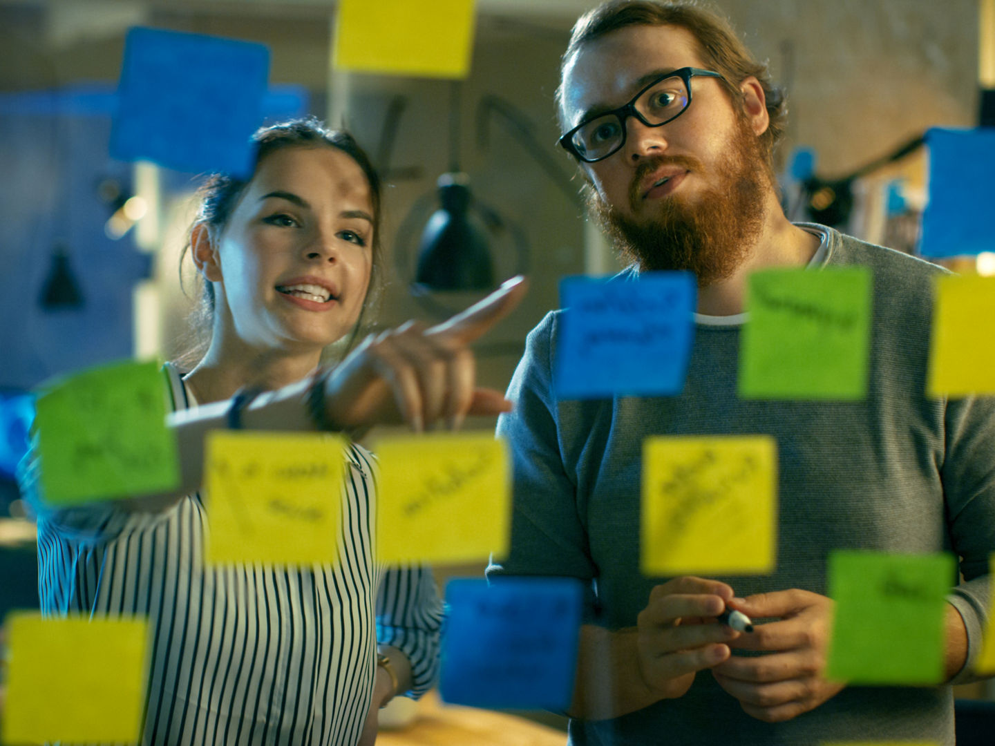 Male and Female Designers Discuss and Outline They're Project with Sticky Notes on a Glass Wall. They're Young and Creative People Who Work in a Stylish Loft Space.