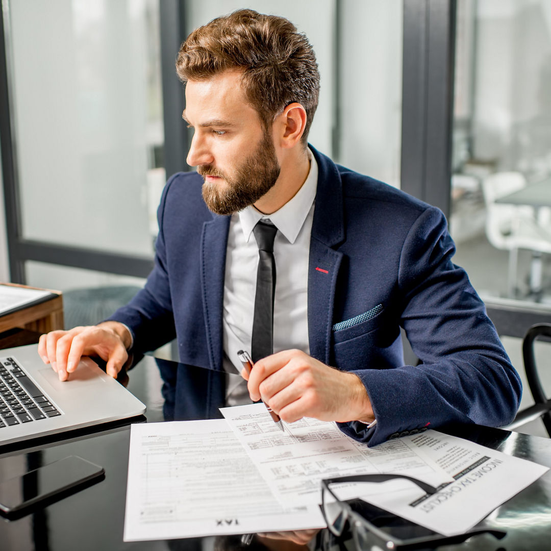 Handsome tax manager dressed in the suit working with documents and laptop at the modern office interior