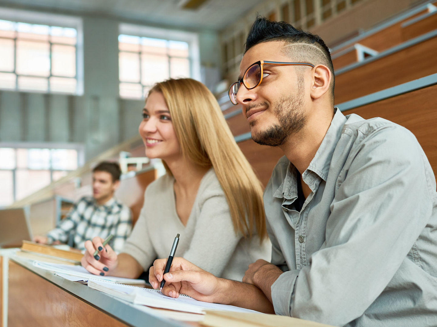 Multi-ethnic group of students sitting at desk in lecture hall of modern college and smiling happily, focus on young Middle-Eastern man wearing glasses, copy space