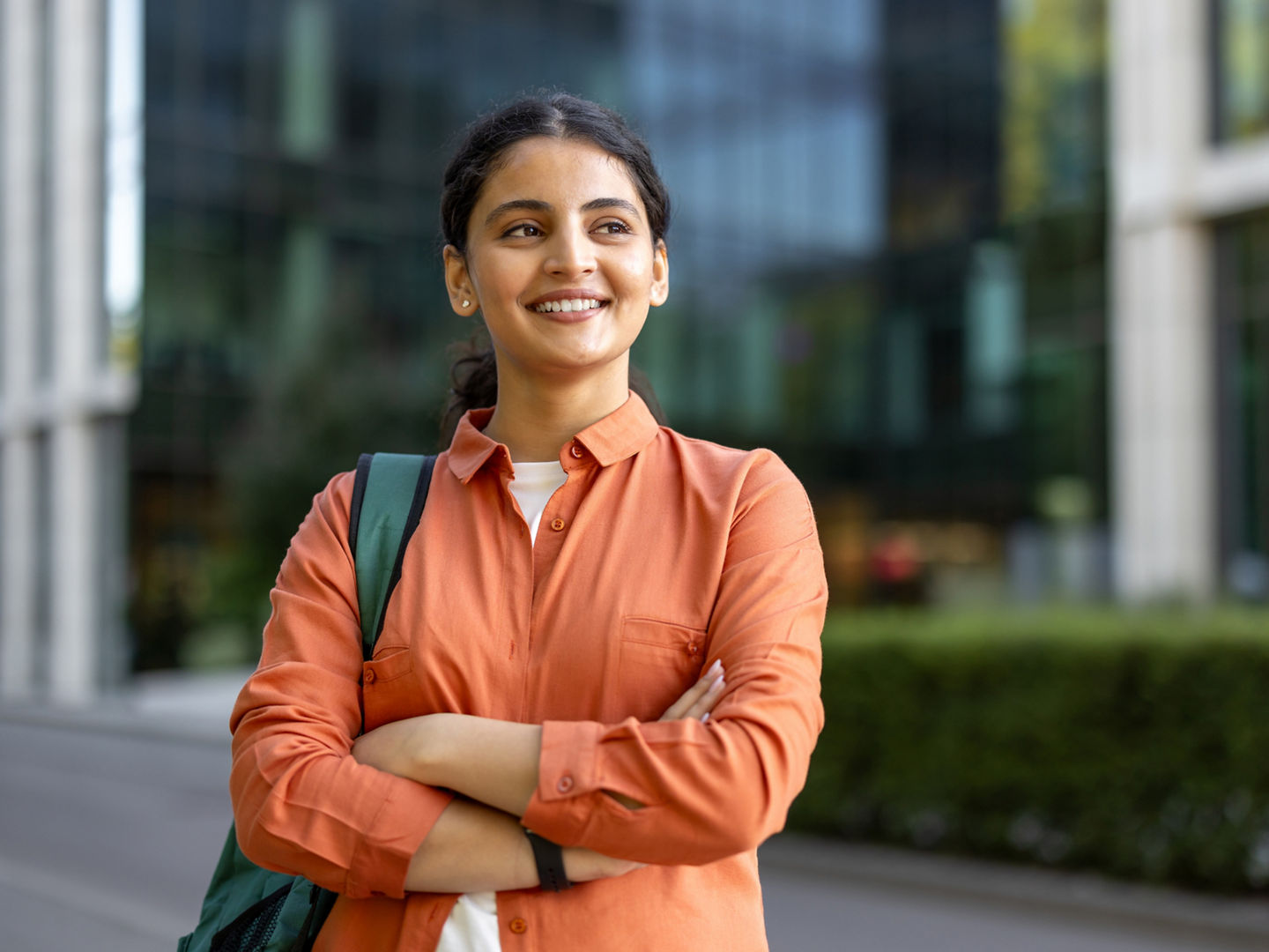 Young indian woman carrying a backpack and crossing arms, looking away and smiling in front of a modern university building, representing education, success, and future opportunities