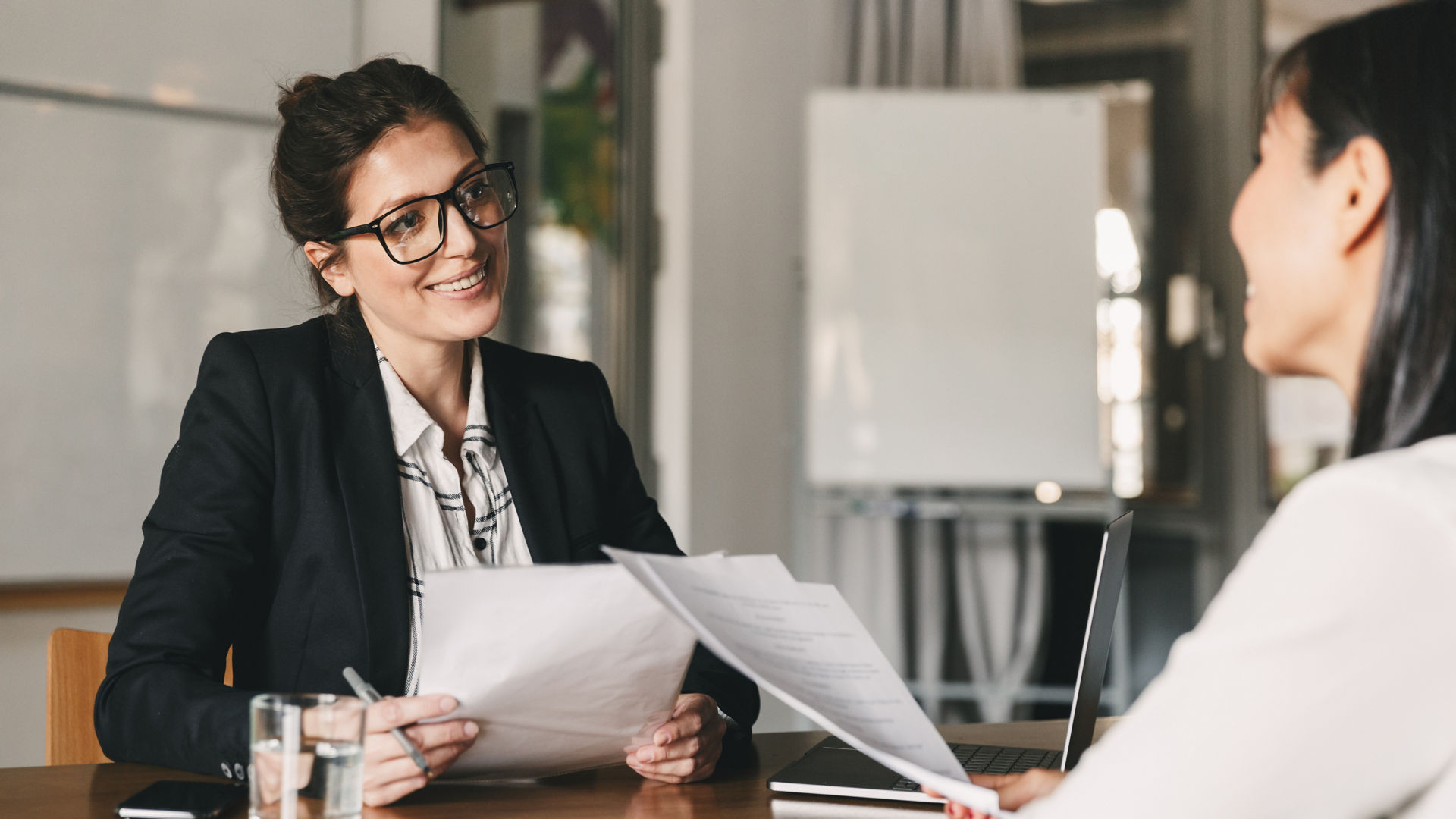 Portrait of smiling businesswoman holding resume and talking to female candidate during corporate meeting or job interview - business, career and placement concept