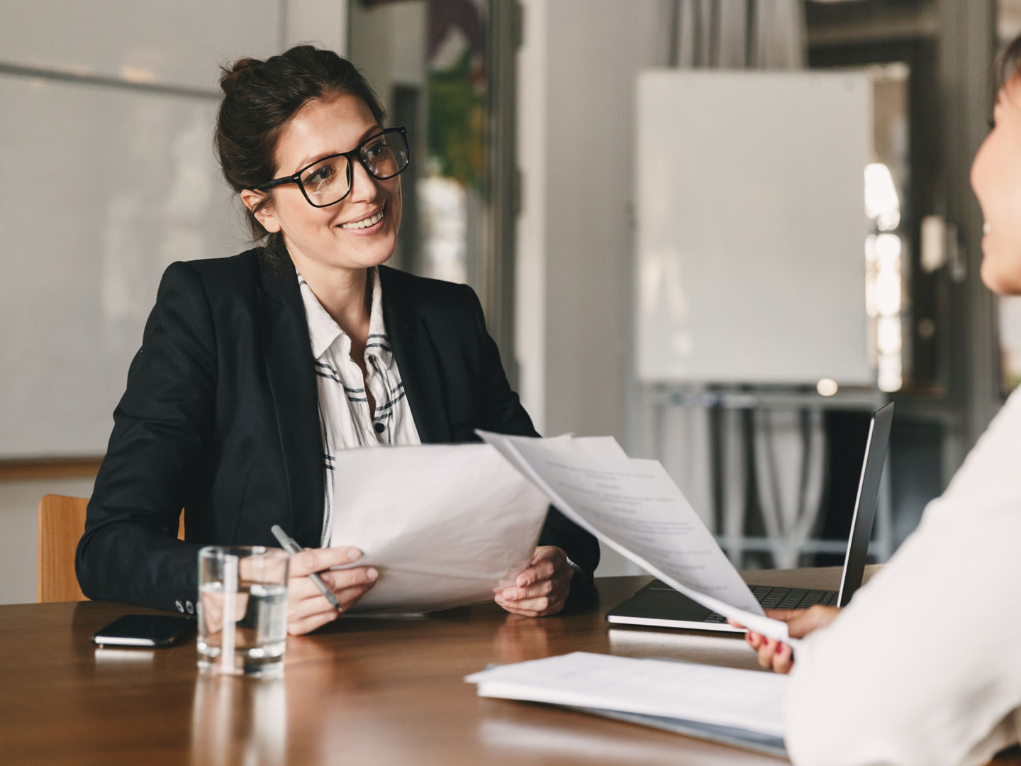 Portrait of smiling businesswoman holding resume and talking to female candidate during corporate meeting or job interview - business, career and placement concept