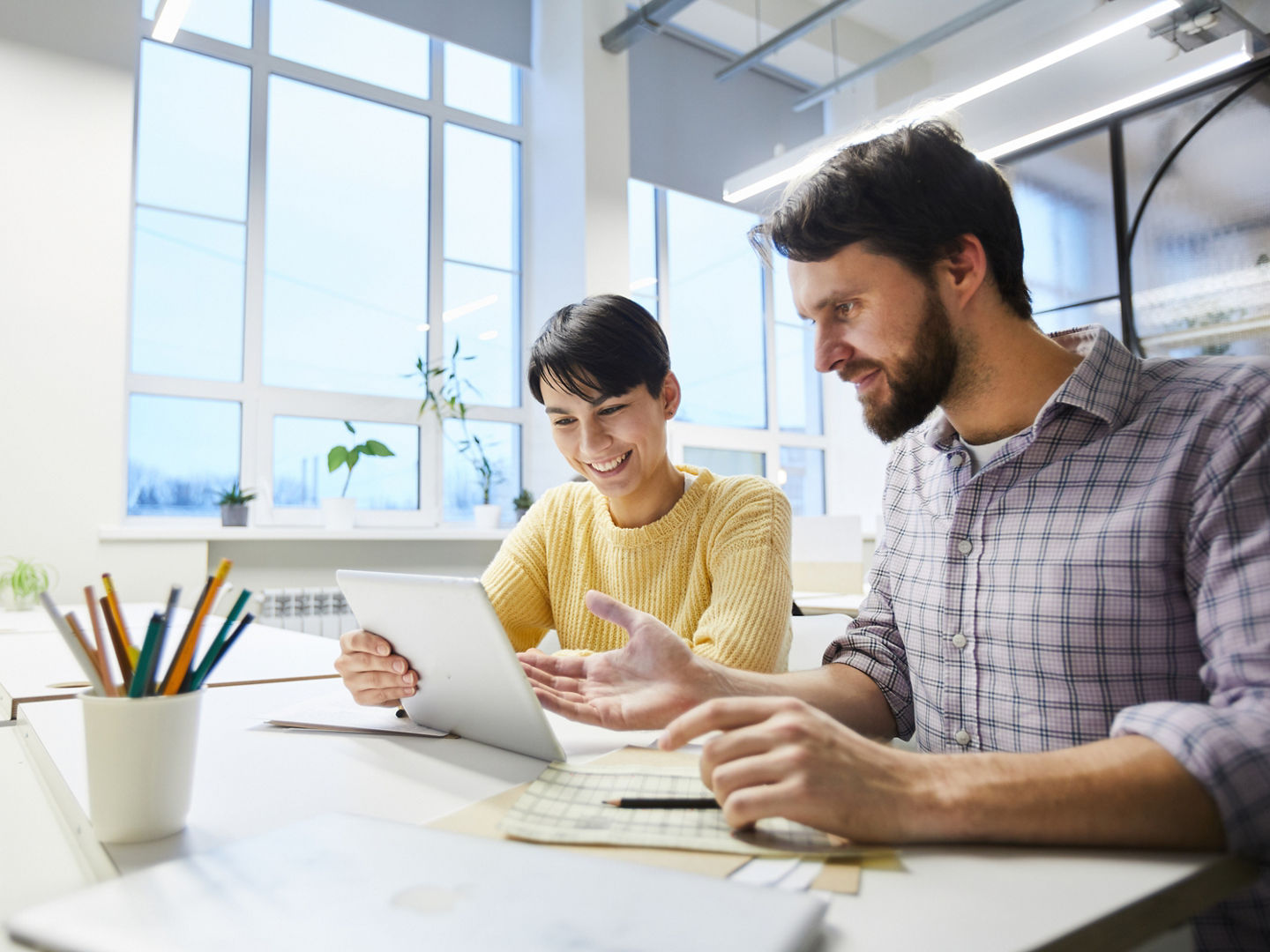 Positive excited young colleagues in casual clothing sitting at table and discussing internet project while using modern tablet in office