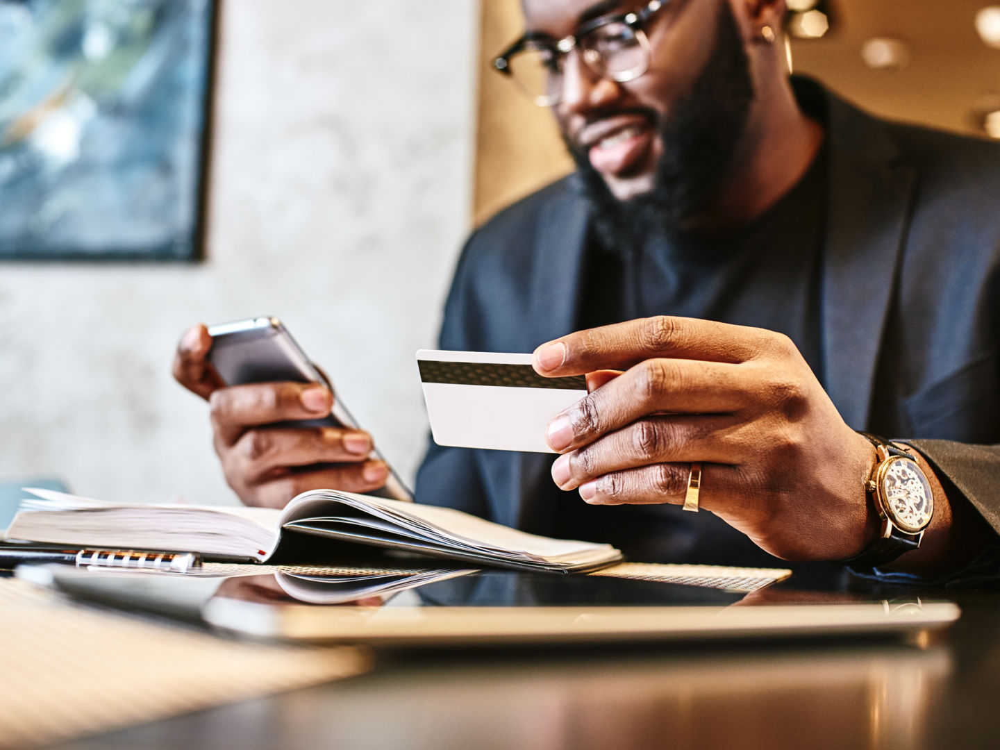 Portrait of African-American male holding cell phone in one hand and credit card in other, making transaction, using mobile banking app during lunch at cafe. His working papers and tablet pc are on the table. Online payment and shopping concept.