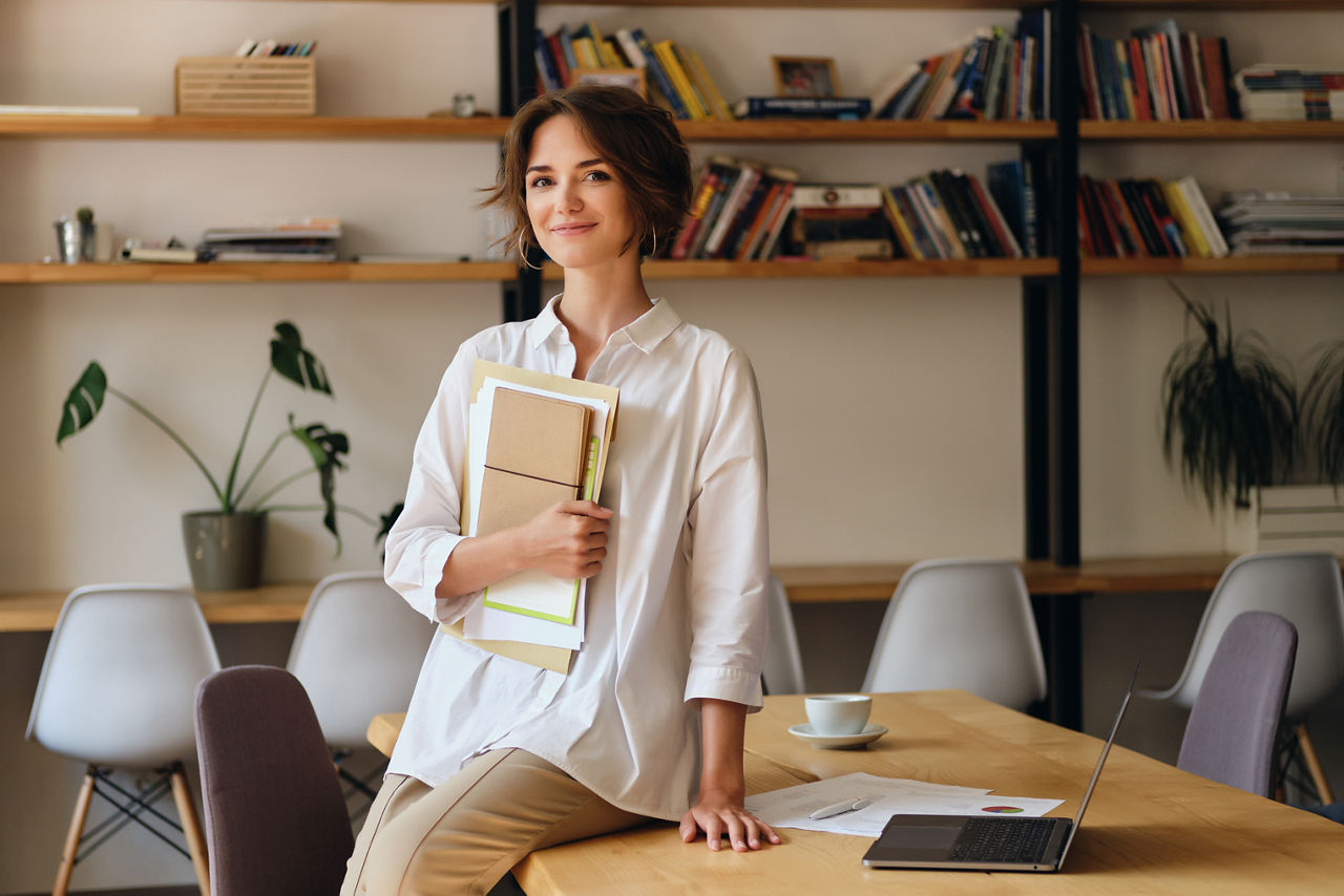 Young attractive woman in white shirt happily looking in camera sitting on desk with papers and laptop in modern office