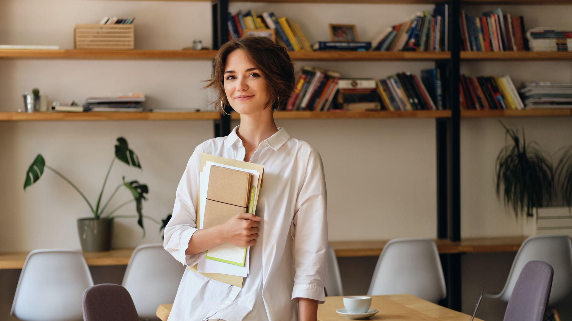 Young attractive woman in white shirt happily looking in camera sitting on desk with papers and laptop in modern office