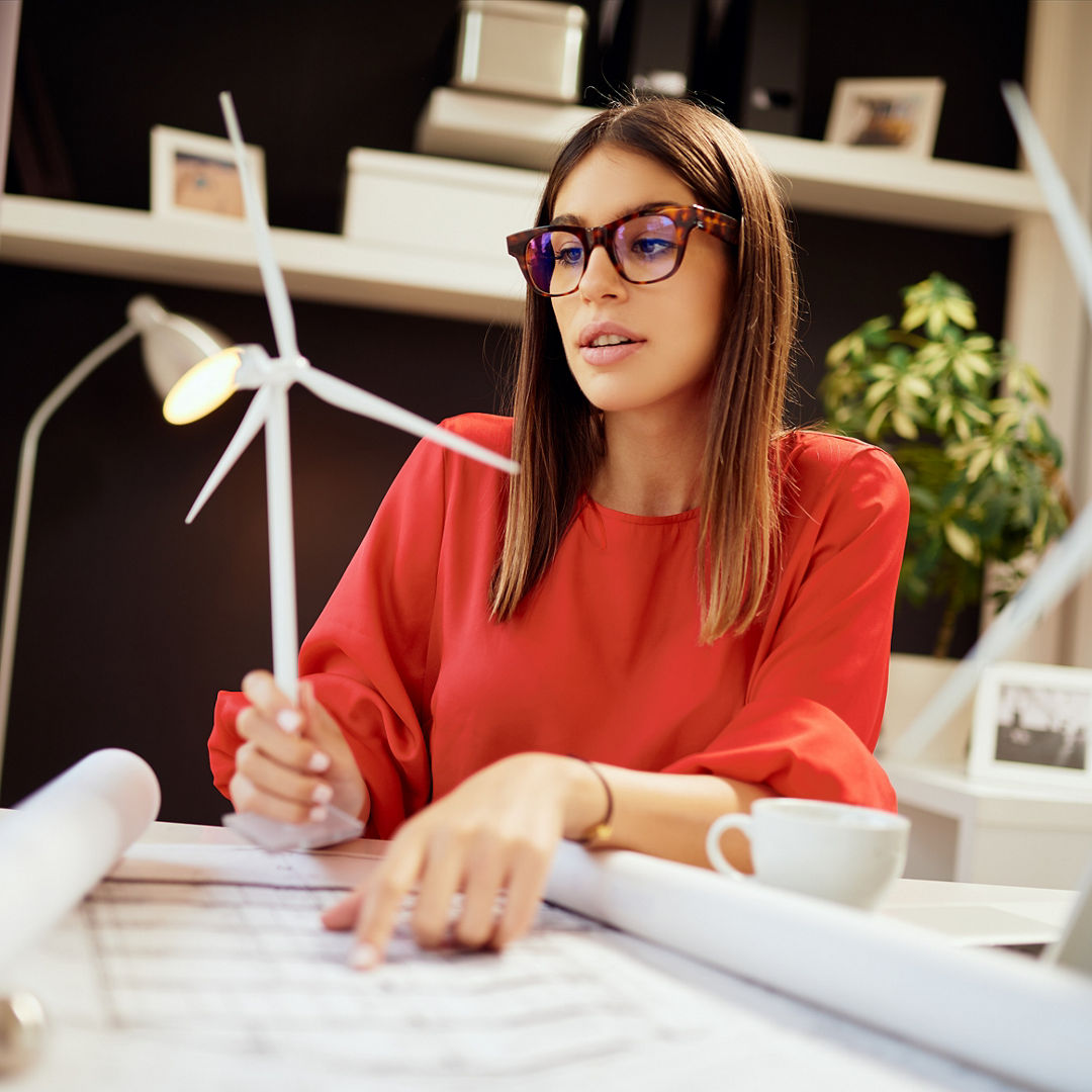 Dedicated hardworking pretty businesswoman dressed in formal wear sitting in office and holding windmill model. On table are blueprints and laptop. Sustainable development concept.