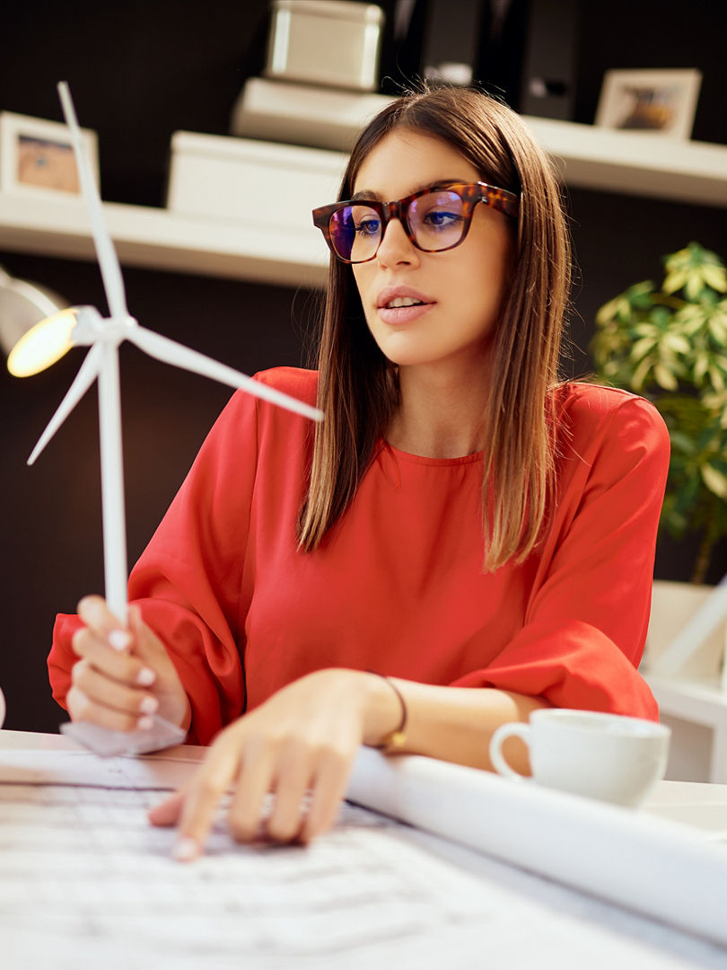 Dedicated hardworking pretty businesswoman dressed in formal wear sitting in office and holding windmill model. On table are blueprints and laptop. Sustainable development concept.