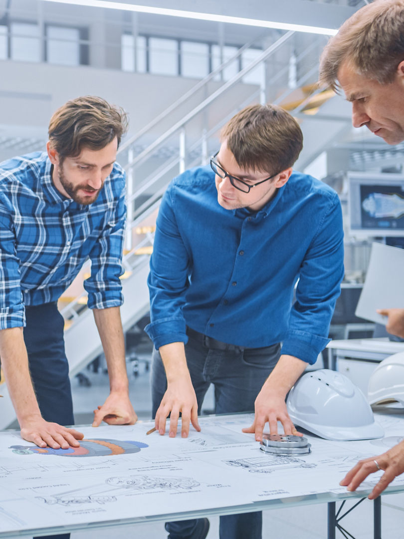In the Industrial Engineering Facility: Diverse Group of Engineers and Technicians on a Meeting Gather Around Table with Engine Design Technical Drafts, Have Discussion, Analyse Technology