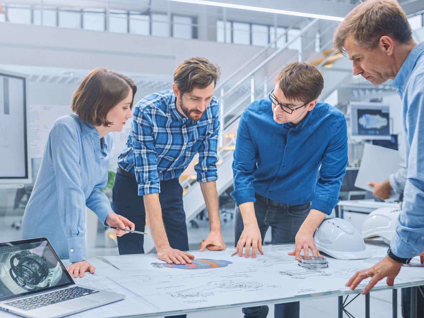 In the Industrial Engineering Facility: Diverse Group of Engineers and Technicians on a Meeting Gather Around Table with Engine Design Technical Drafts, Have Discussion, Analyse Technology