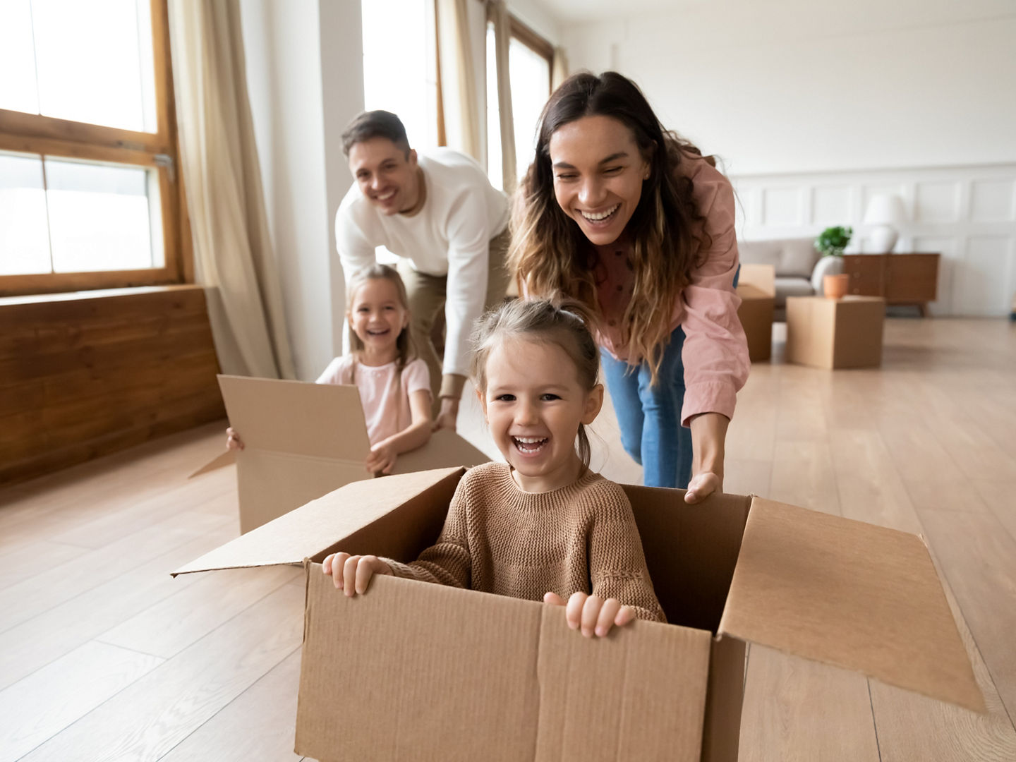 Happy parents playing with cute small kids daughters laughing on moving day, family tenants renters homeowners and children girls having fun riding in box in living room relocating new home concept