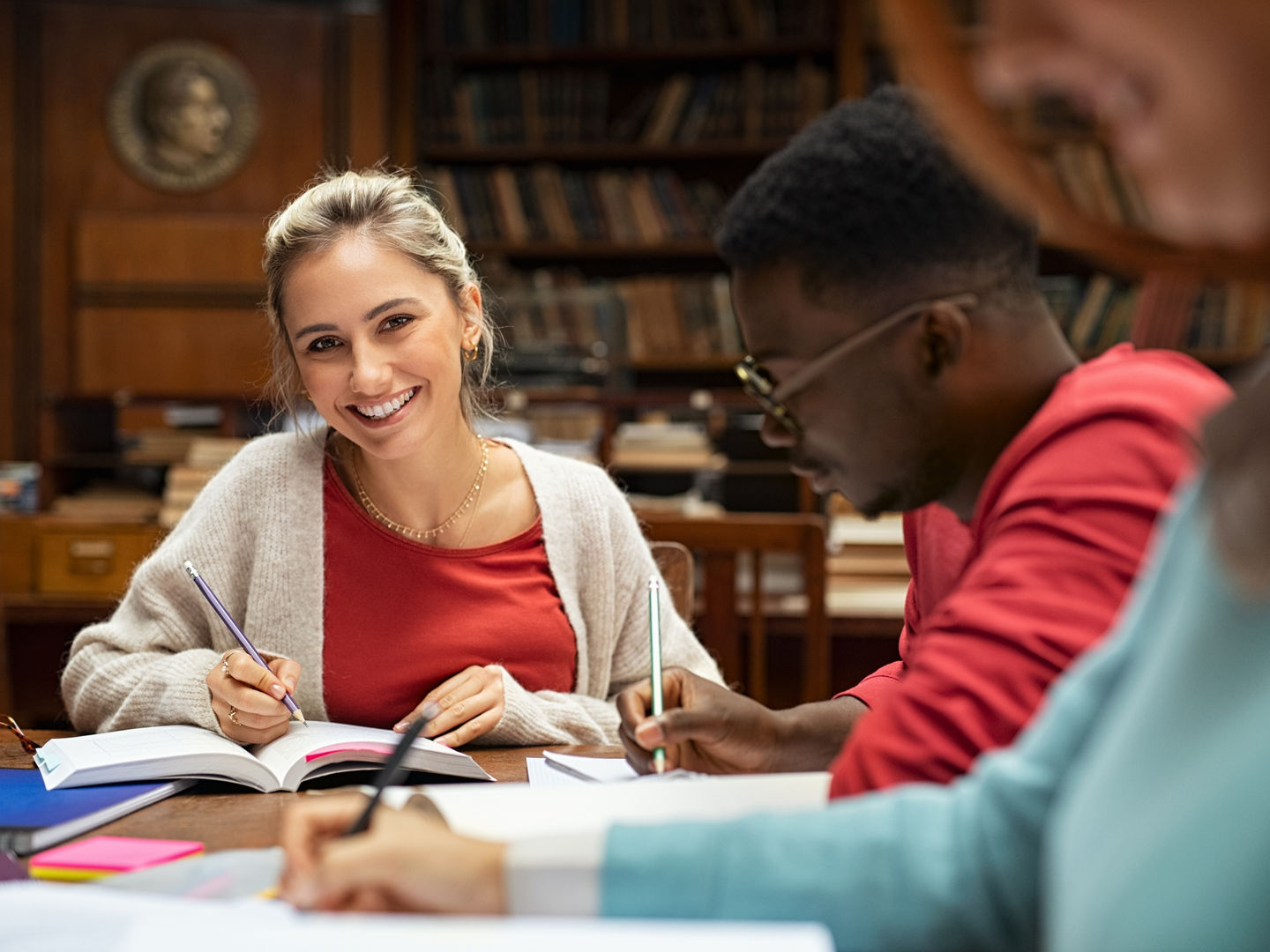 Young woman studying at the library with her classmates. Portrait of beautiful girl at the university smiling and looking at camera. Satisfied college student studying with friend.