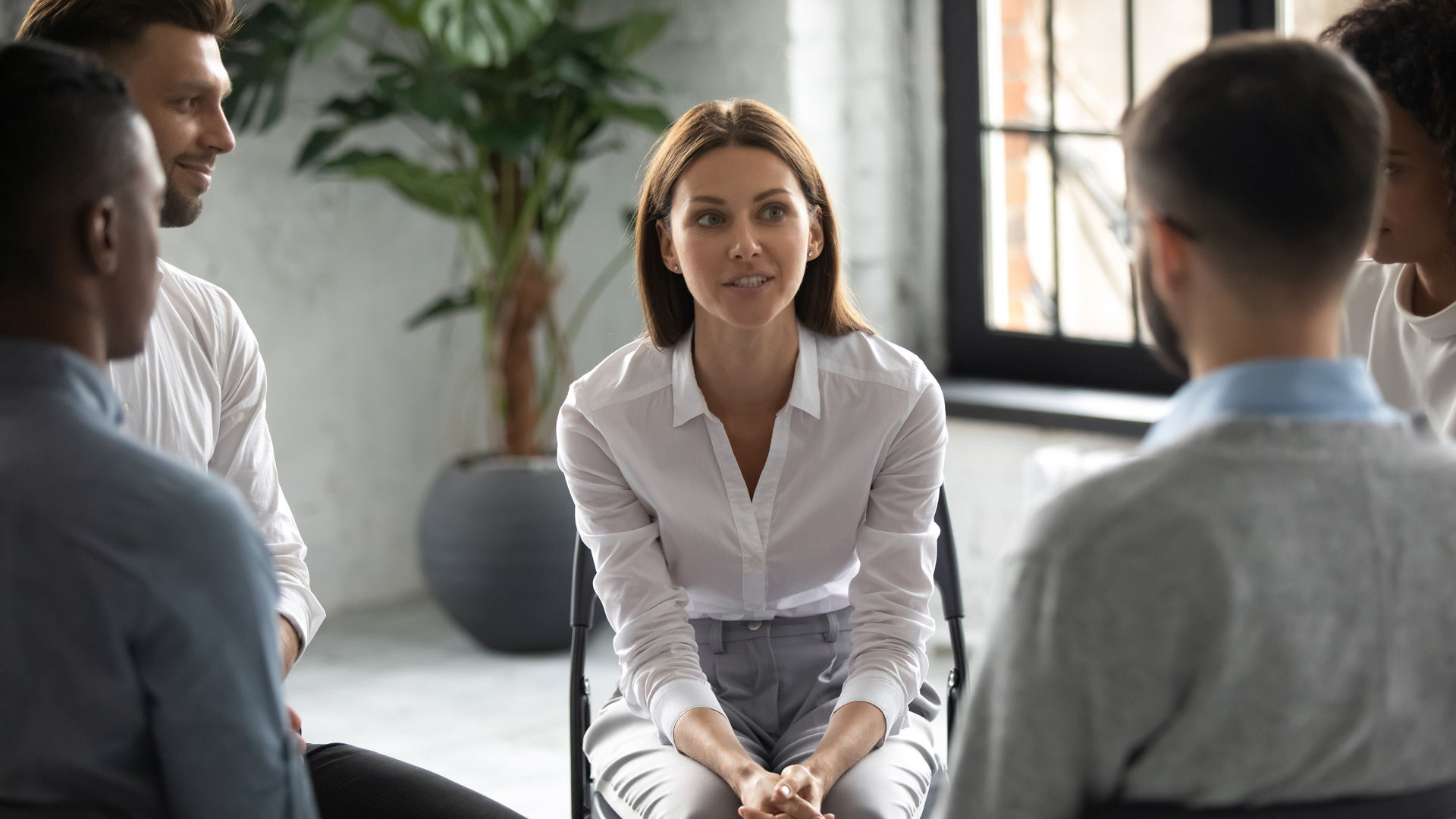 Smiling attractive female psychologist counselling speaking with diverse people sitting in circle at group therapy session. Business coach training staff, having fun, team building activity at work.