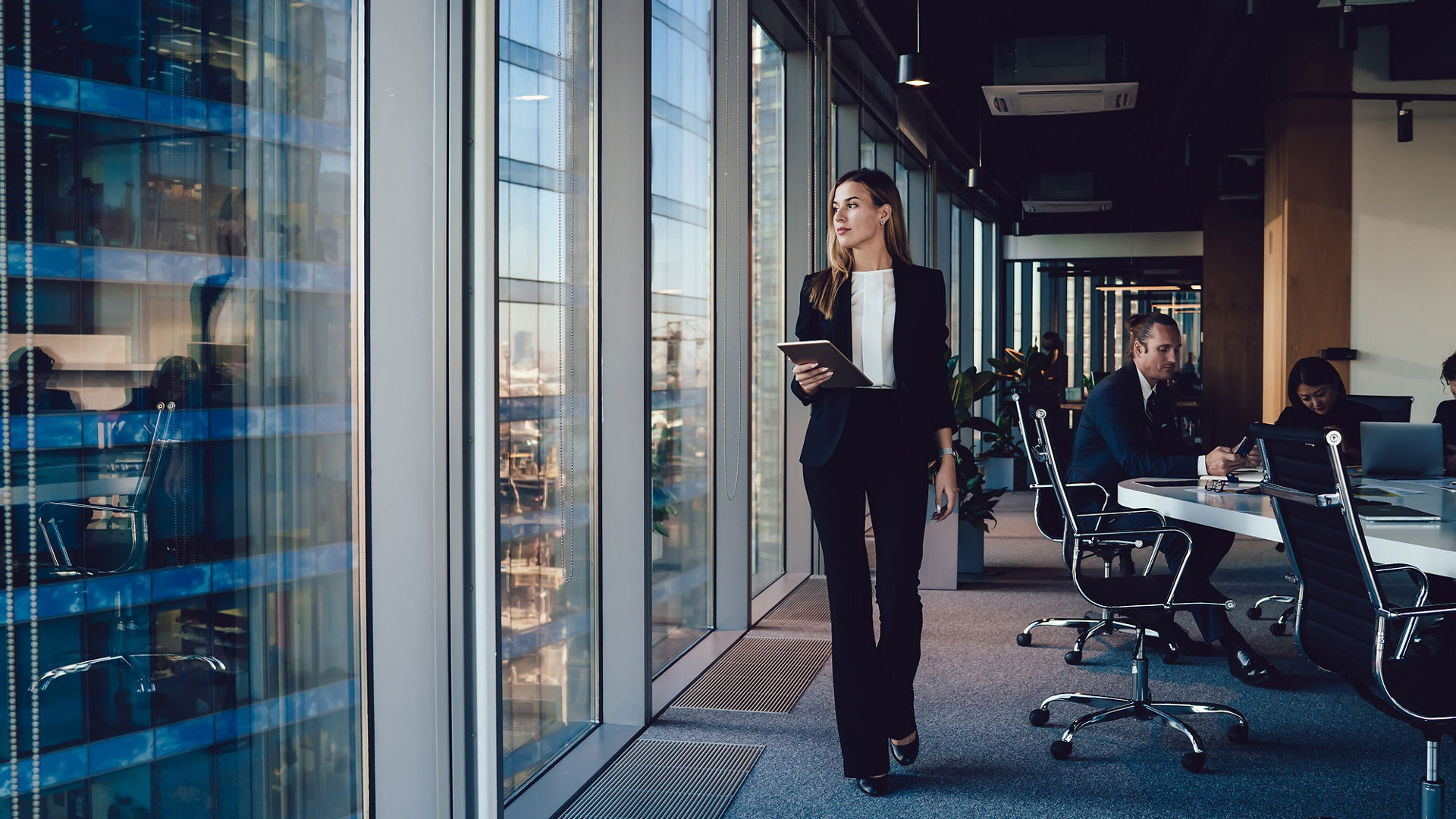 Serious female administrative manager in formal suit walking in office with digital tablet for checking office working process, confident woman business owner looking at window passing with touchpad