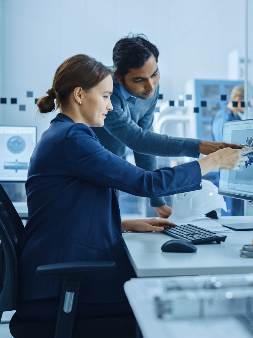 Inside Factory Office: Male Project Supervisor Talks to a Female Industrial Engineer who Works on Computer, Talk. In Workshop: Professional Workers Use High-Tech Industry 4 CNC Machinery, Robot Arm.