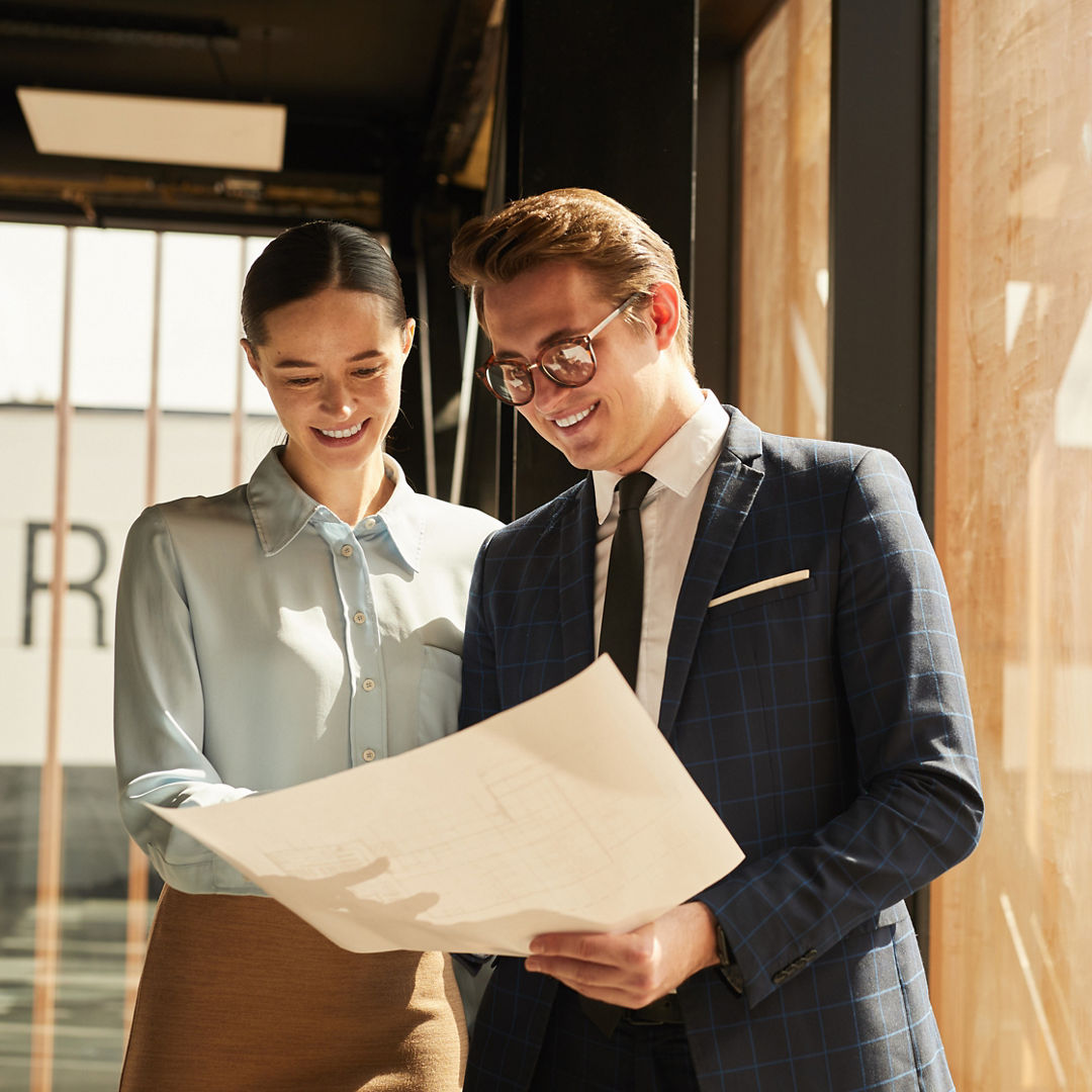Waist up portrait of smiling rental agent showing floor plans to female client while standing in office building interior lit by sunlight, copy space