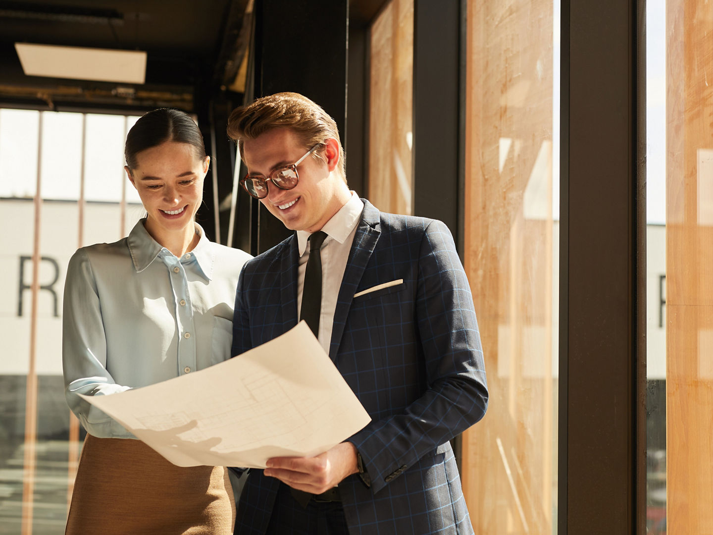 Waist up portrait of smiling rental agent showing floor plans to female client while standing in office building interior lit by sunlight, copy space