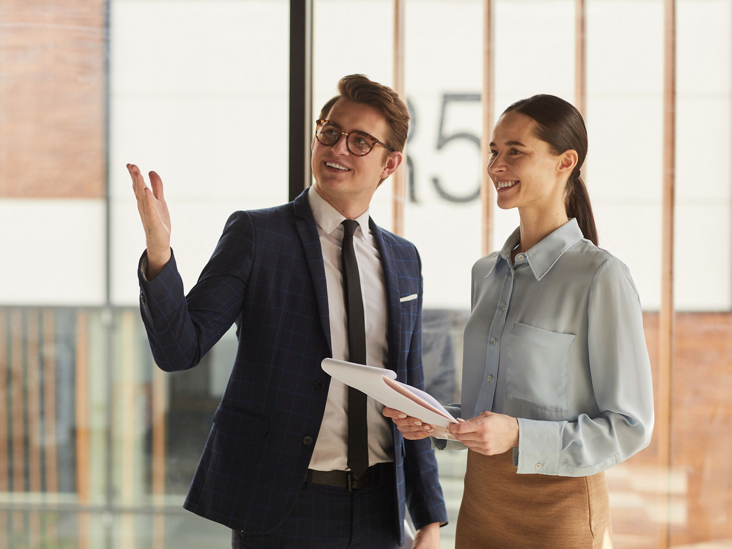 Waist up portrait of smiling real estate agent discussing property with female client and pointing up while standing in empty office building interior lit by sunlight, copy space