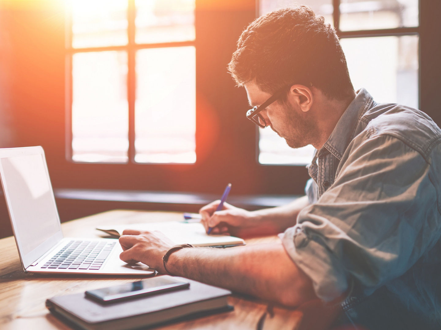 Young millennial male student searching information for homework using laptop and wifi connection indoors, man writing article in textbook for add interesting content on own website or web page