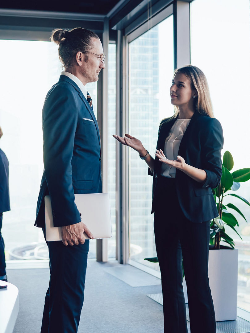Side view of coworking man and woman in formal clothing talking about new project during meeting with colleagues while standing in modern office and looking at each other