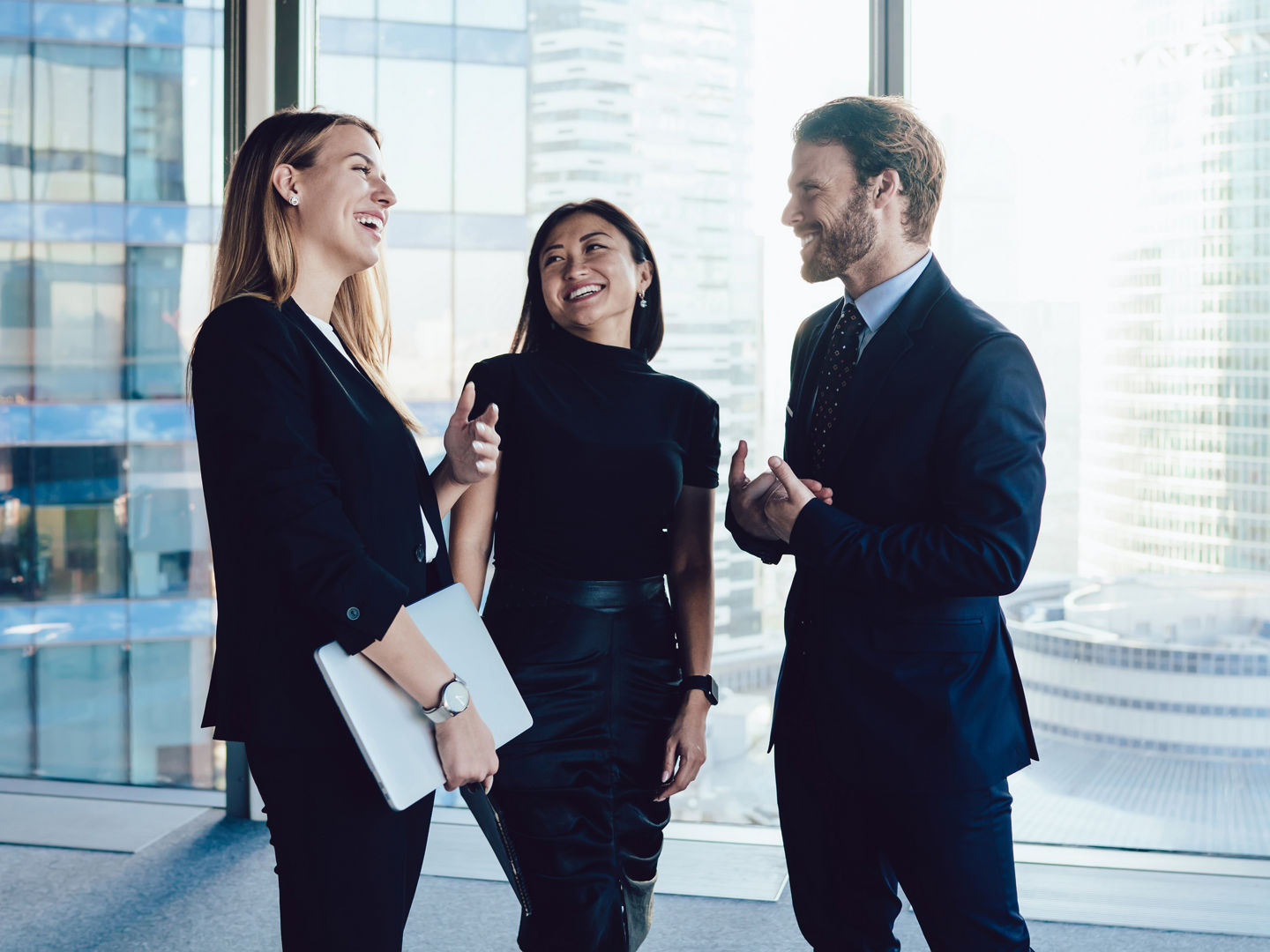 Delighted team of coworkers in formal wear standing in modern corridor and speaking about project while smiling and looking at each other