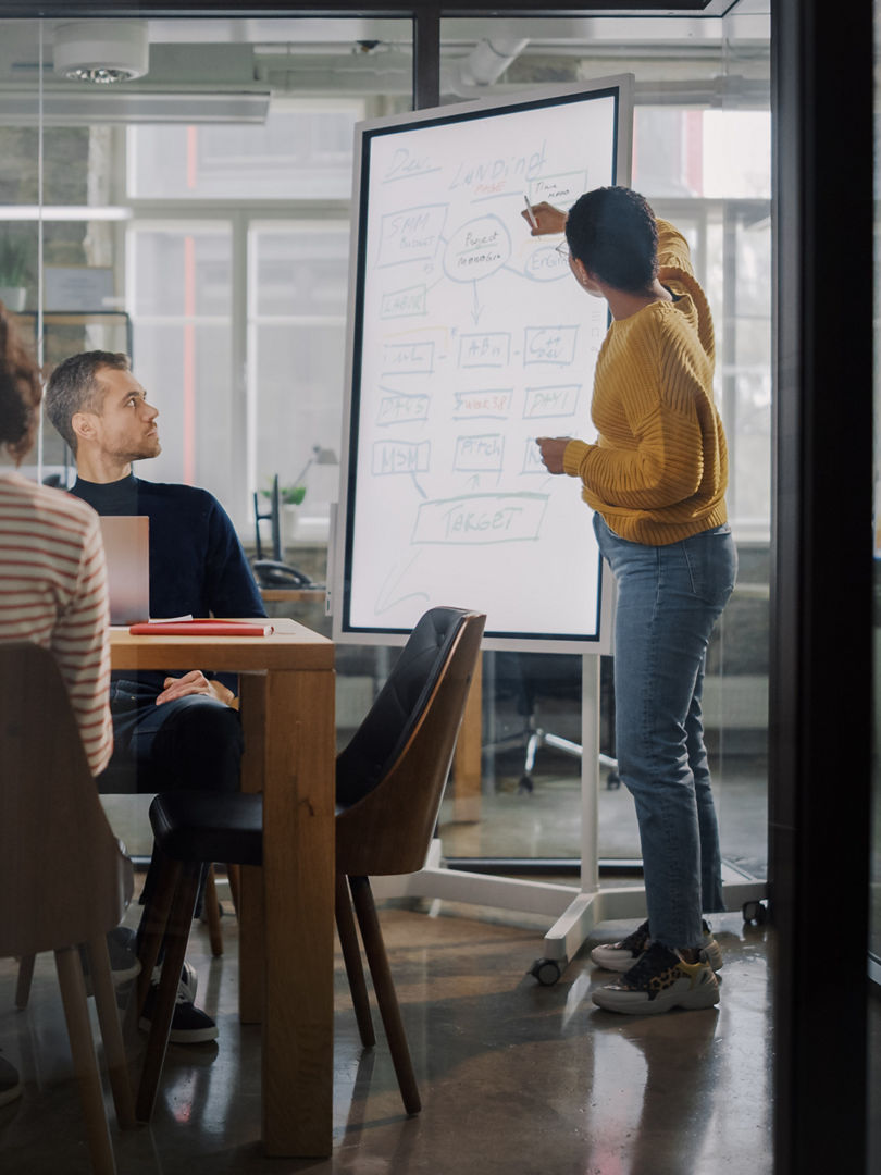 Project Manager Makes a Presentation for a Young Diverse Creative Team in Meeting Room in an Agency. Colleagues Sit Behind Conference Table and Discuss Business Development, User Interface and Design.