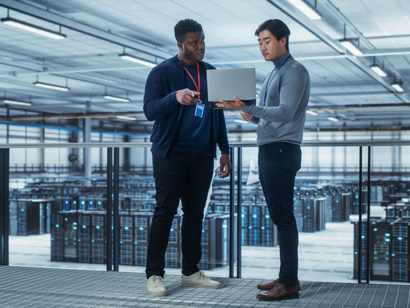 Two Multiethnic Male Data Center System Administrators and IT Specialists Talk, Use Laptop. Information Technology Engineers work on Cyber Security Network Protection in Cloud Computing Server Farm.