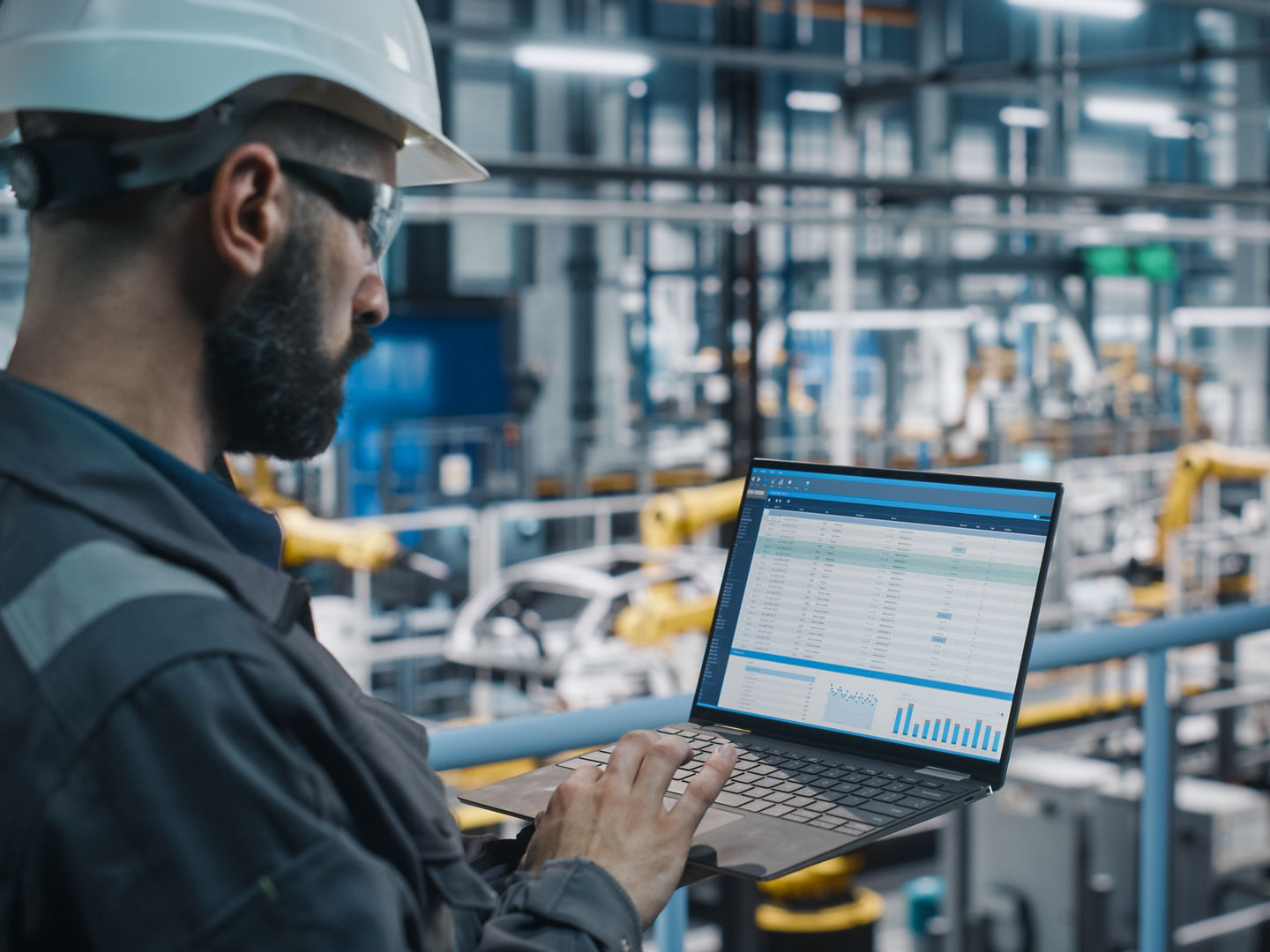 Car Factory Engineer in Work Uniform Using Laptop Computer with Spreadsheet Software. Working with Software at Automotive Industrial Manufacturing Facility Dedicated for Vehicle Production.