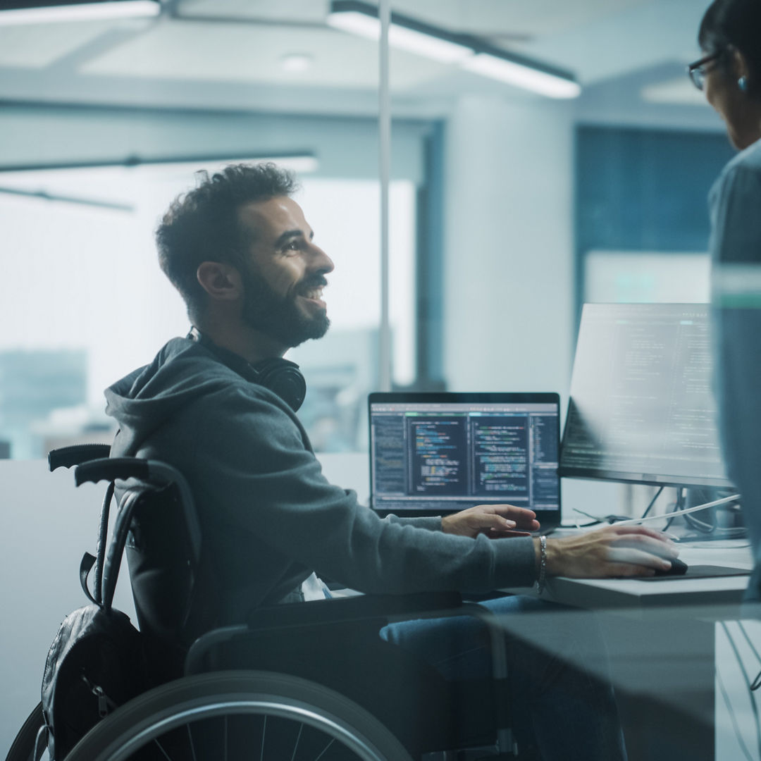 Disability-Friendly Office: Brilliant IT Programmer with Disability in a Wheelchair Talks with Colleague while Working on Computer. Software Engineers Develop Innovative App, Program.