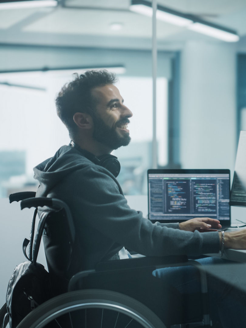 Disability-Friendly Office: Brilliant IT Programmer with Disability in a Wheelchair Talks with Colleague while Working on Computer. Software Engineers Develop Innovative App, Program.