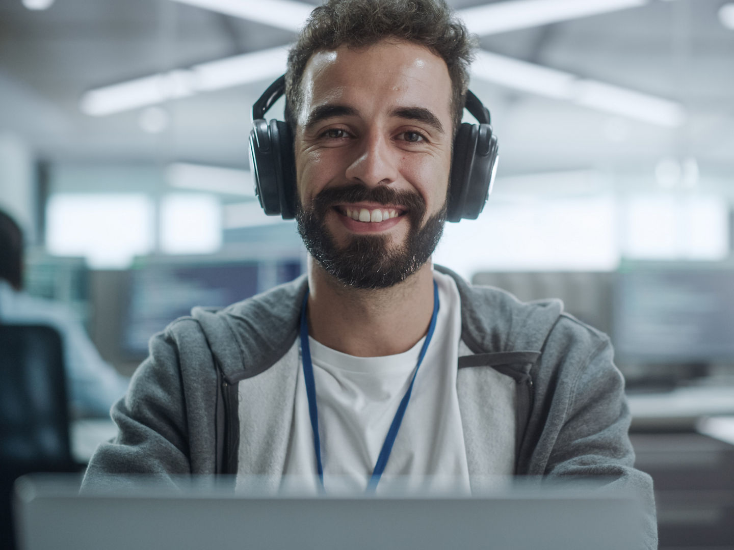 Office: Portrait of Happy IT Programmer Wearing Headphones Working on Desktop Computer, Looking at Camera and Smiling. Male Software Engineer Developing App, Video Game. Listening to Podcast, Music