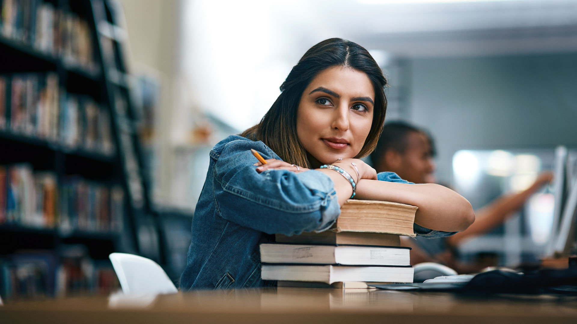 Shot of a young woman resting on a pile of books in a college library and looking thoughtful.