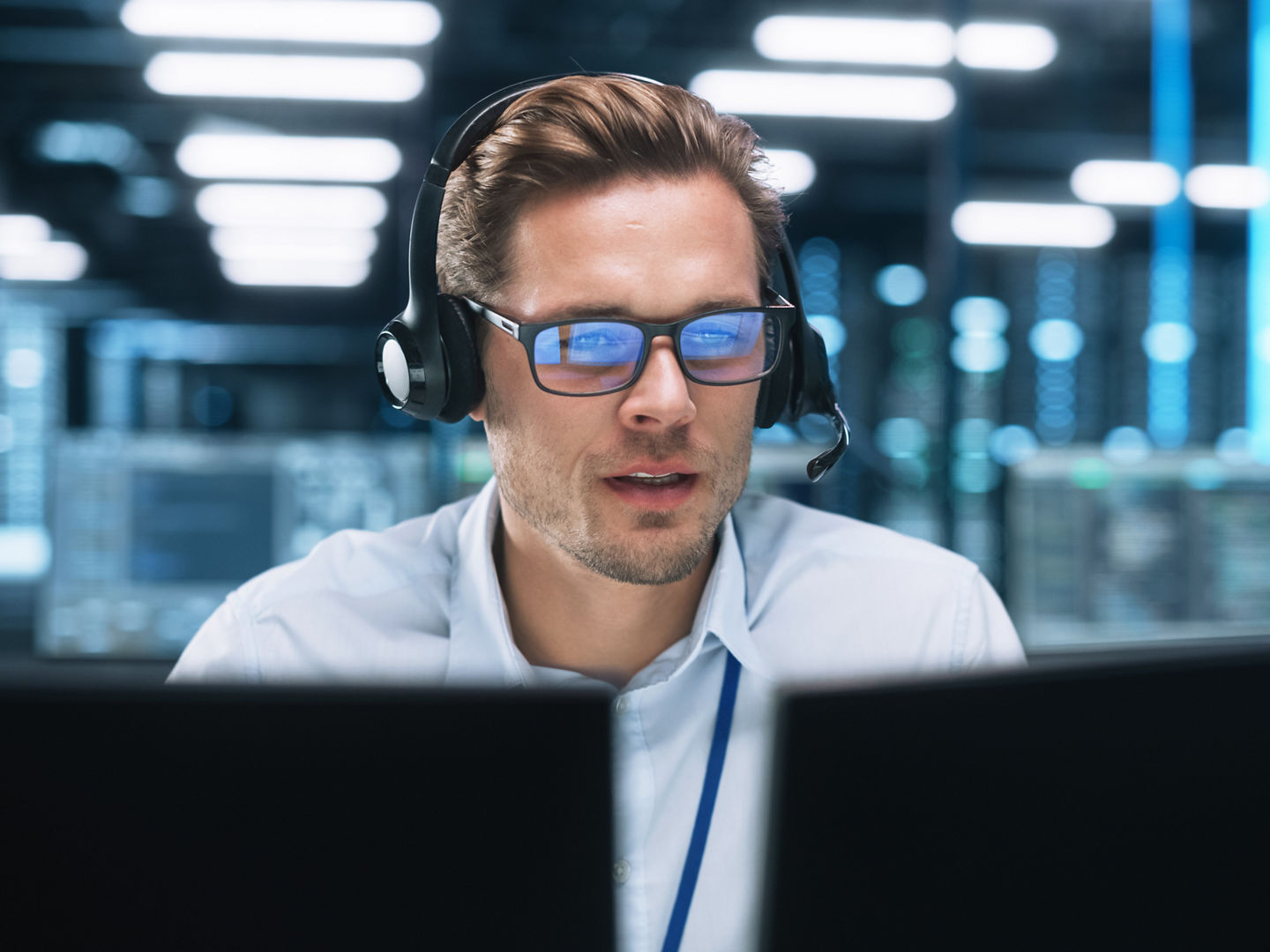 Portrait of a Young Caucasian Male Call Centre Worker Wearing a Phone Headset Talking at the Support Line with Serious Face in a Modern Open Plan Office at Night. Helpline Concept