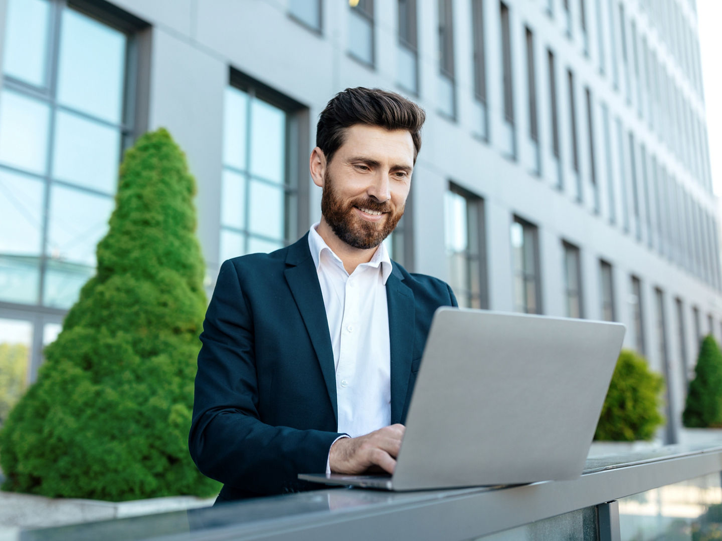 Smiling confident millennial attractive european businessman with beard in suit chatting on laptop near modern office building. Manager work outdoor, successful business with technology and new normal