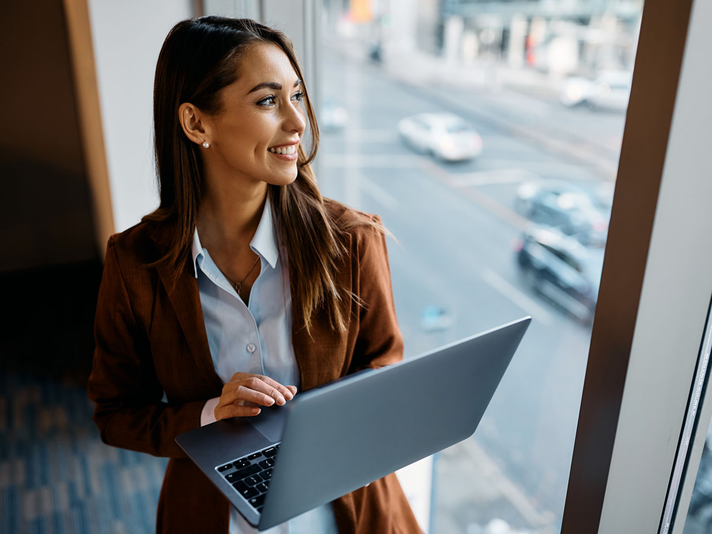 Happy female entrepreneur using laptop and looking through window in office.