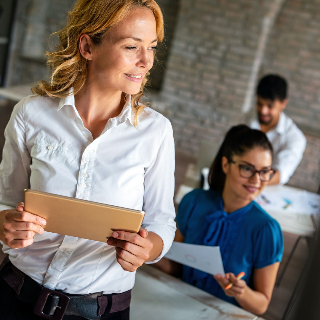 Young successful attractive female business manager working on digital tablet while standing in modern office with colleagues in background
