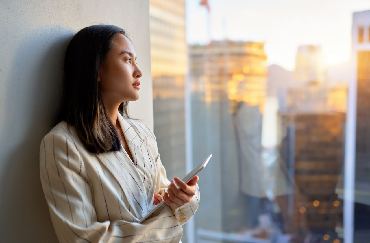 Young pensive Asian business woman professional manager executive leader holding digital tablet standing in corporate office looking at window with city view, thinking of solution, lost in thought.