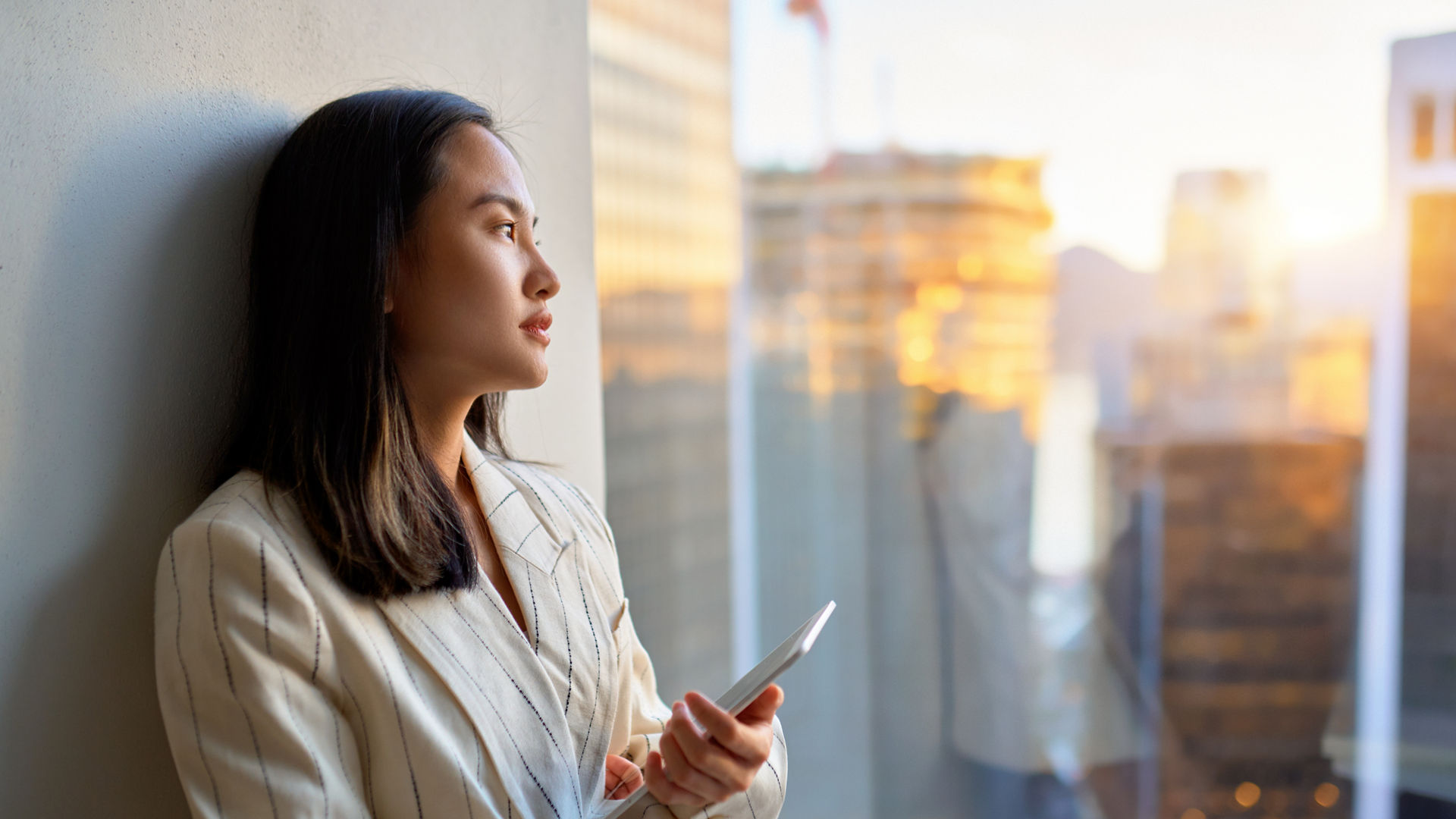 Young pensive Asian business woman professional manager executive leader holding digital tablet standing in corporate office looking at window with city view, thinking of solution, lost in thought.