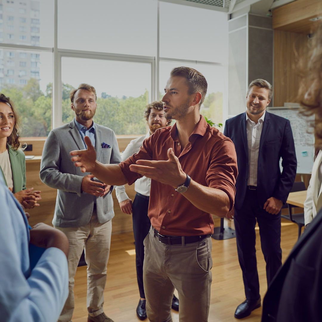 Experienced business coach meeting with a group of people. Young male business trainer or psychologist standing in a circle of men and women, talking about teamwork and giving professional advice