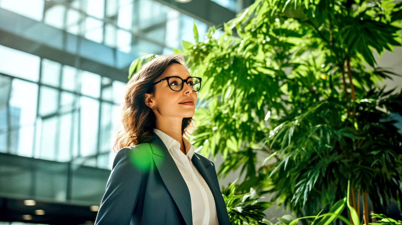 Female startup entrepreneur stands amidst vibrant green foliage in modern office hall. Concept of investment on corporate sustainability and modern technology solutions. Banner. Copy space.