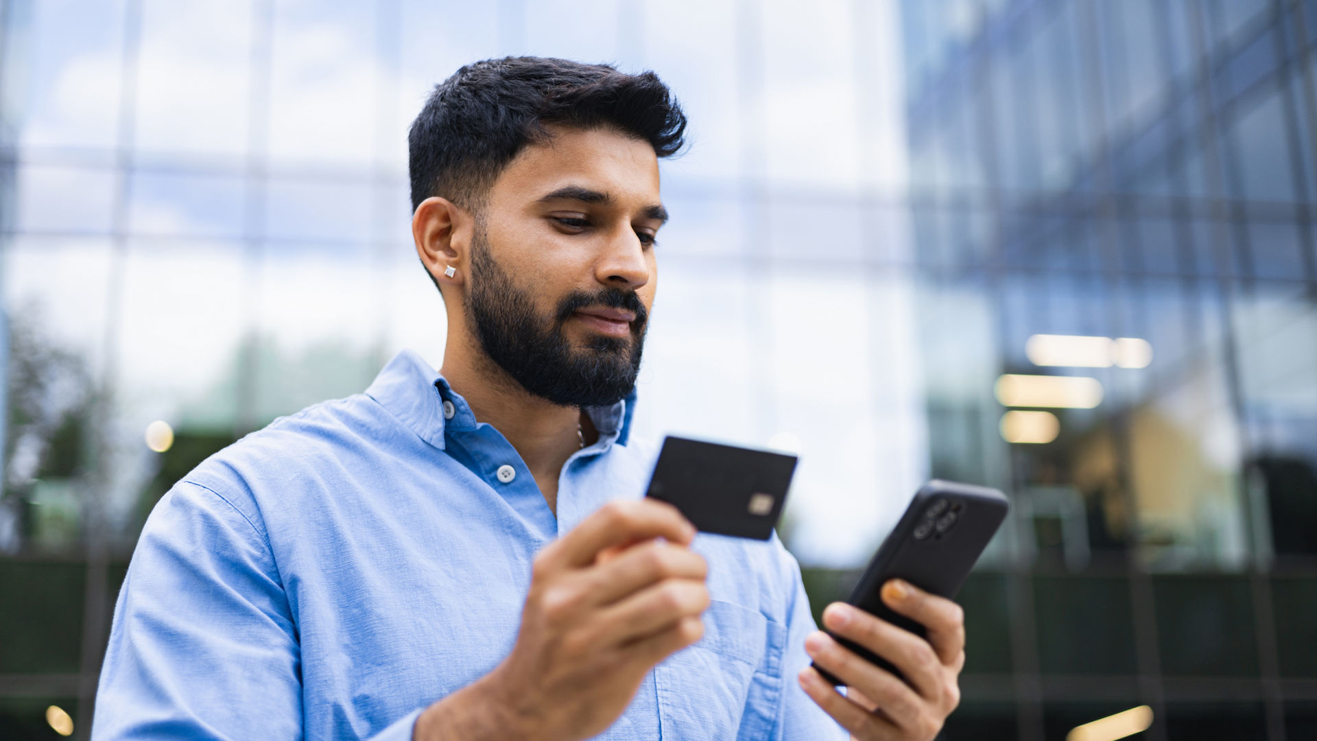 An Indian man in a blue shirt is shown holding a credit card and a smartphone in front of a modern building.