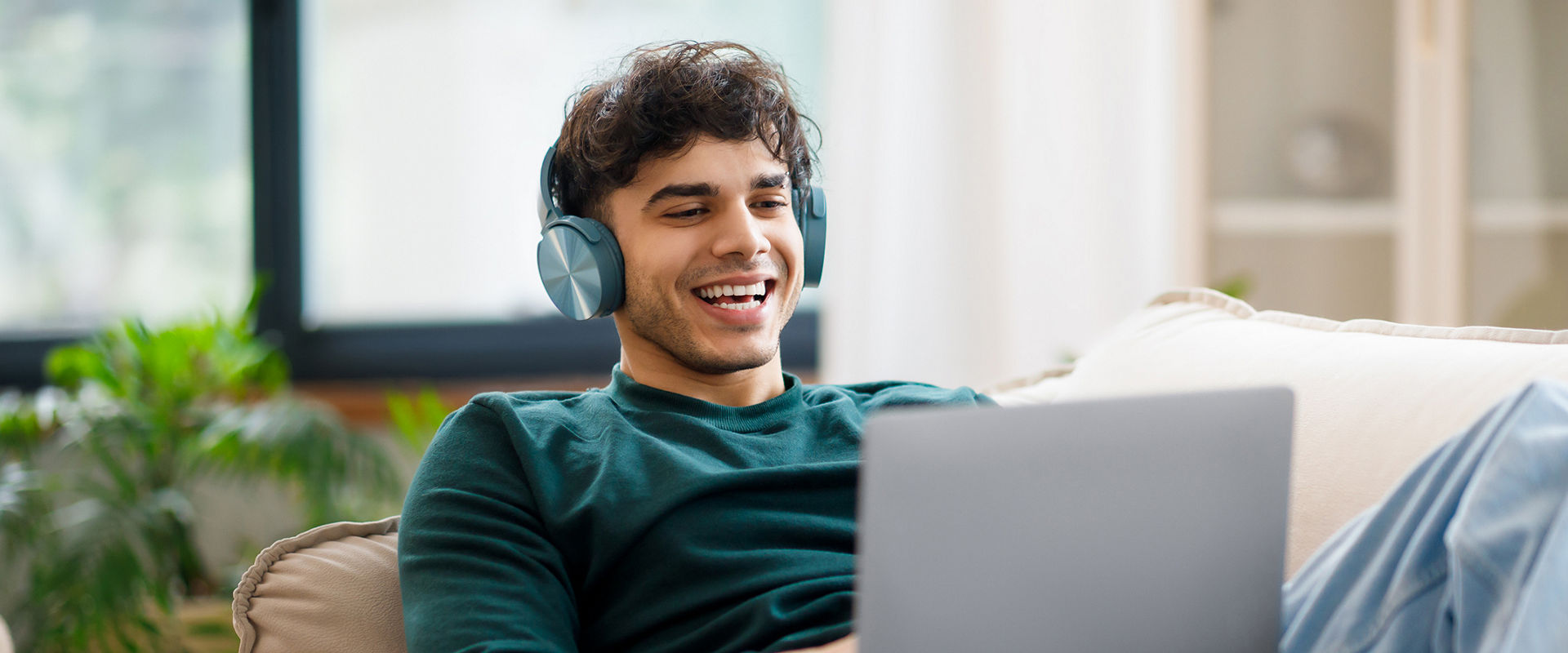 Online Fun. Smiling Arab Guy In Wireless Headphones Using Laptop At Home, Relaxing On Couch Watching Movie On Computer Or Listening To Music. Freelancer Enjoying Remote Job