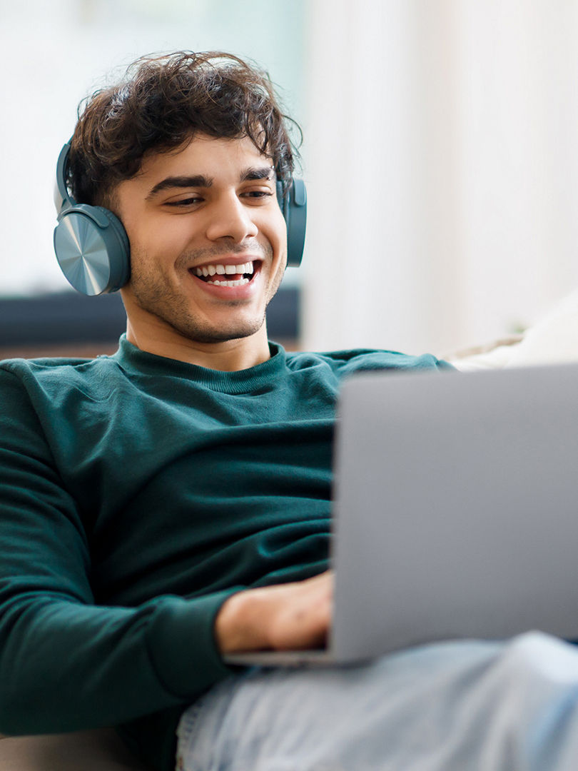 Online Fun. Smiling Arab Guy In Wireless Headphones Using Laptop At Home, Relaxing On Couch Watching Movie On Computer Or Listening To Music. Freelancer Enjoying Remote Job