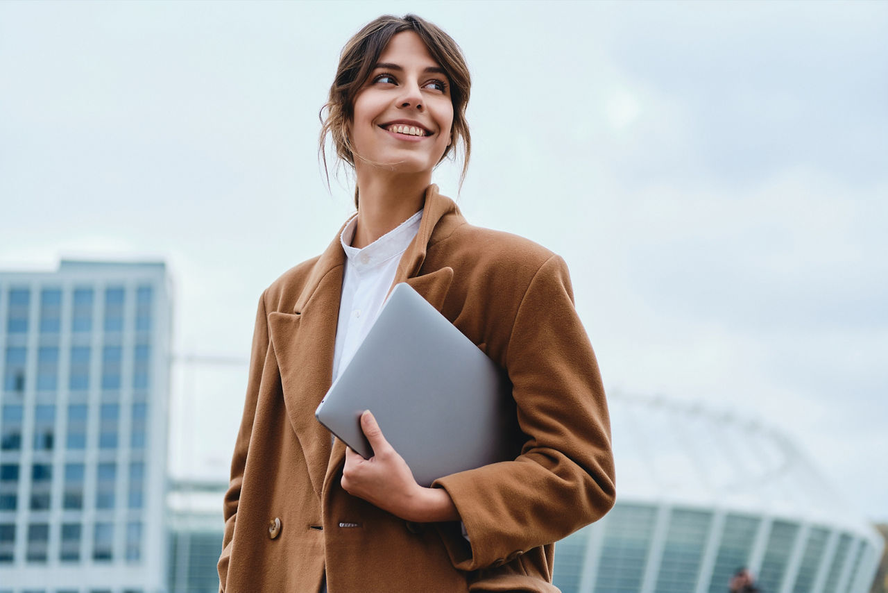 Young pretty smiling businesswoman in coat with laptop happily looking away on city street